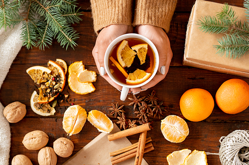 Top view of aromatic tea with orange slices held by female surrounded by fresh fruit, spices and walnuts on wooden table