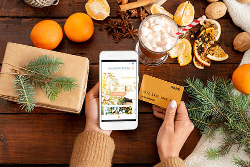 Overview of human hands with smartphone and credit card surrounded by Christmas objects over wooden table