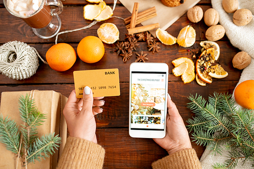 Top view of human hands with plastic card and smartphone over wooden table with Christmas gift, conifer and fruit