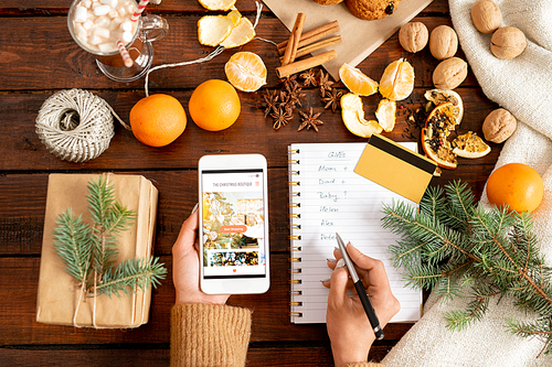 Female hands holding smartphone and making list of Christmas gifts to buy in online shop before holiday