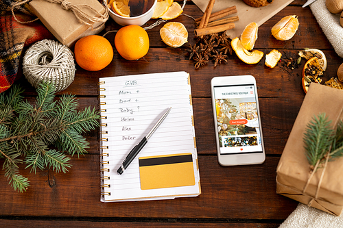 View of smartphone with promo, notepad with pen, list and credit card surrounded by traditional xmas stuff on table