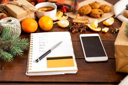 Christmas composition of smartphone, notepad page with list, pen and credit card among festive stuff on wooden table