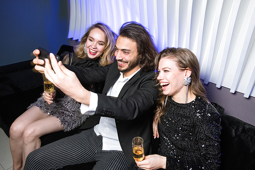 Happy young glamorous women with champagne and elegant man with smartphone making selfie on couch in night club