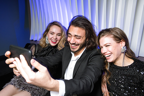 Laughing glamorous girls and elegant man with smartphone making selfie while enjoying party in night club