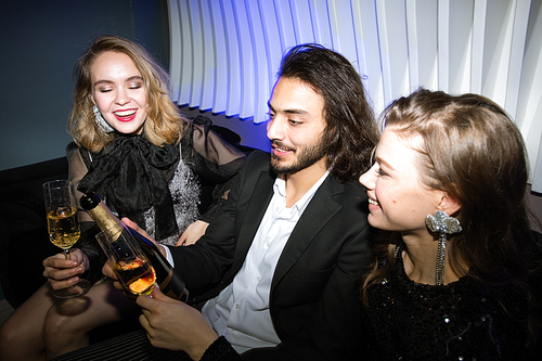 Happy glamorous girls with flutes of champagne and young man holding bottle while sitting on couch in night club and enjoying party
