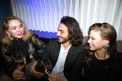 Happy gorgeous girls and young elegant man toasting with champagne in night club while enjoying party