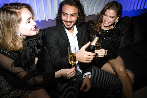 Happy girls and young elegant man sitting on couch in night club and toasting with champagne at party