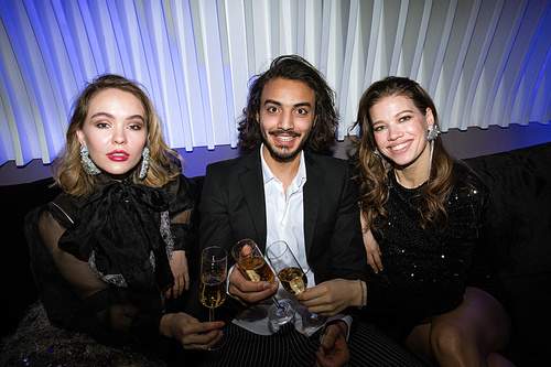 Three young glamorous friends with flutes of champagne sitting on couch in night club, toasting and enjoying party