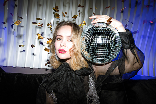 Pretty young blond female in black evening dress holding glittering disco ball while enjoying party in night club
