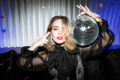 Pretty blond girl in black organza evening dress holding glittering disco ball by her head while enjoying party in night club
