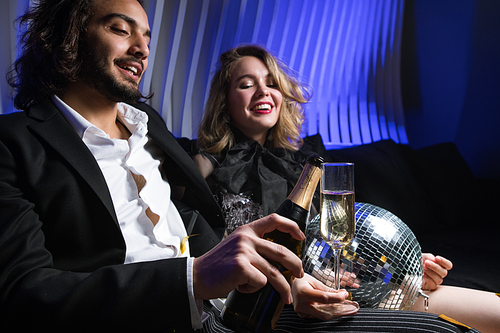 Happy young man with bottle of champagne going to pour wine into flute of his girlfriend while relaxing at party