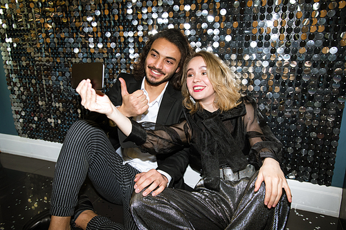 Cheerful young well-dressed couple making selfie while sitting by glittering wall in night club at party