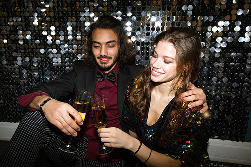 Young affectionate couple clinking with flutes of champagne at party in night club by glittering wall