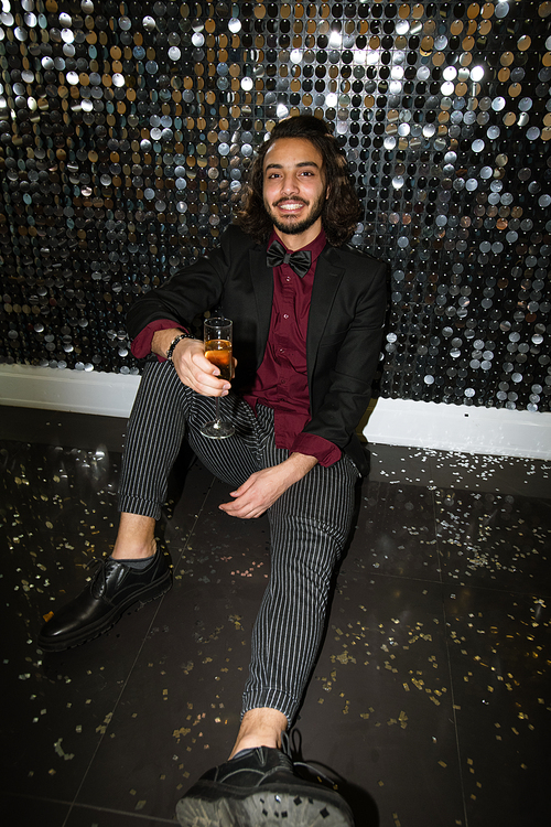 Happy young elegant man in suit toasting with flute of champagne while sitting on the floor by glittering wall at party