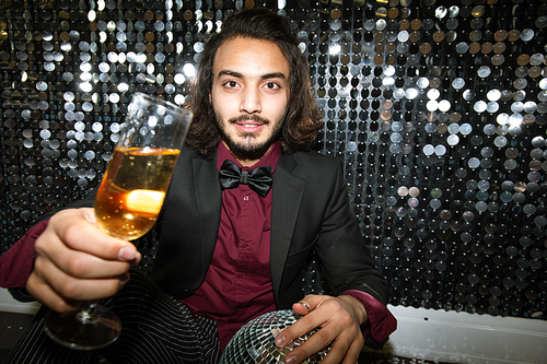 Happy young well-dressed man cheering up with flute of champagne while sitting on the floor by glittering wall at party