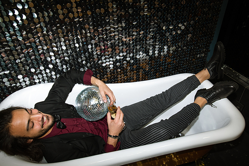 Cheerful young well-dressed man with flute of champagne relaxing in bathtub at party in the night club