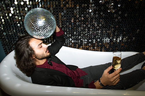Young tired man with disco ball by head and flute of champagne relaxing in bathtub at party in the night club