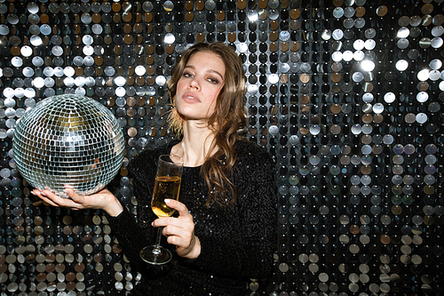 Pretty young woman with flute of champagne and disco ball looking at you while toasting at party in the night club