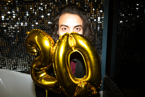 Happy young brunette male looking at you while holding golden inflatable balloon numbers by his face at party