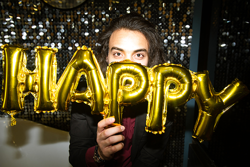 Young brunette male looking at you while holding golden inflatable balloon letters by his face at party in the night club