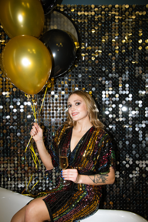 Happy young glamorous woman with flute of champagne and bunch of balloons sitting by glittering wall at party