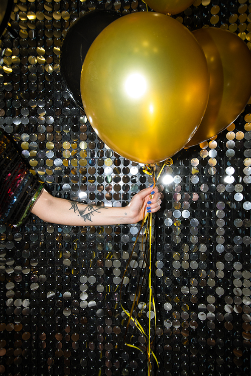 Hand of young woman with tattoo holding golden balloon against glittering wall at party in the night club