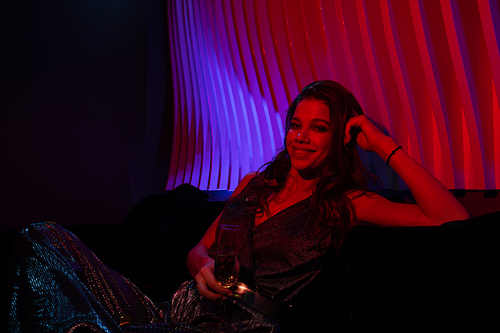 Pretty young woman in glittering attire having champagne while relaxing on couch in dark room at party
