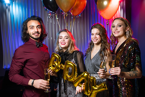 Three happy girls and young man holding flutes of champagne and balloons while celebrating birthday in the night club