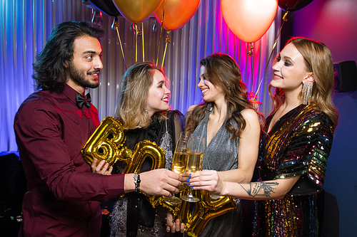 Happy young man clinking flute of champagne with one of girls at birthday party on background of two female friends