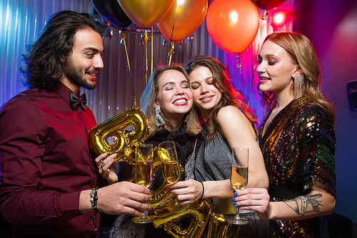 Two young smiling women clinking flutes of champagne with elegant man while enjoying birthday party in the night club