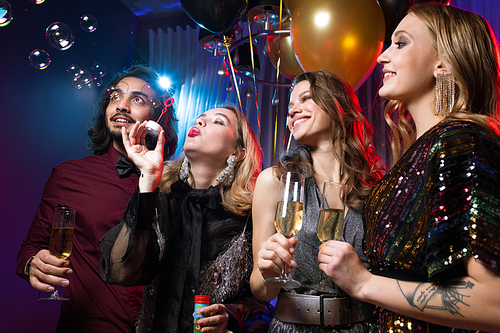 Glamorous girl blowing soap bubbles among friends with flutes of champagne at birthday party