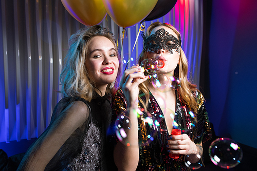 One of two pretty girls blowing soap bubbles with her friend standing near by at party in the night club