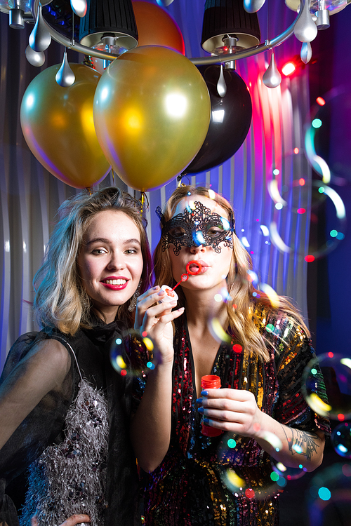 Pretty girl in lace venetian mask blowing soap bubbles with her friend standing near by at party in the night club