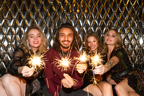 Three happy girls and guy with sparkling bengal lights sitting by wall in the night club in front of camera