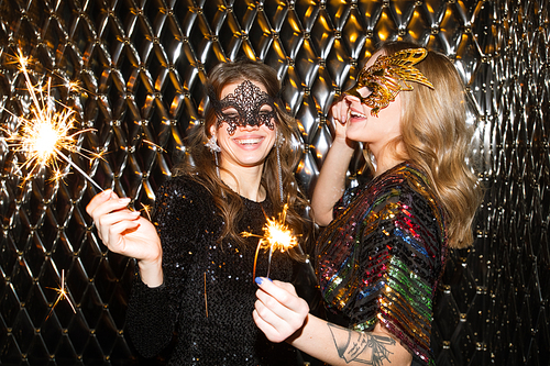 Two joyful girls in venetian masks holding sparkling bengal lights while having fun at party in the night club