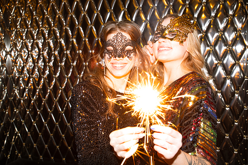 Two cheerful girls in venetian masks holding sparkling bengal lights while enjoying party in the night club