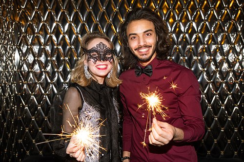 Happy girl in venetian mask and guy with sparkling bengal lights standing against wall in the night club in front of camera