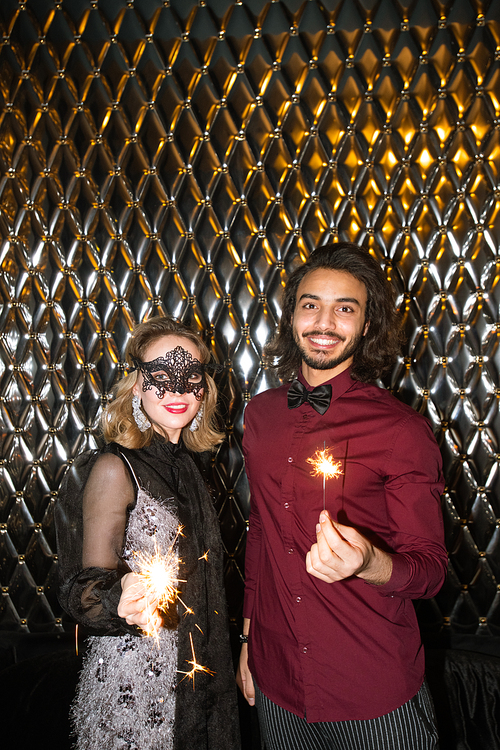 Glamorous girl in venetian mask and elegant guy with sparkling bengal lights standing against wall in front of camera