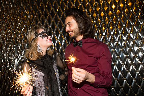 Smiling girl in venetian mask and her boyfriend holding sparkling bengal lights while looking at each other against wall