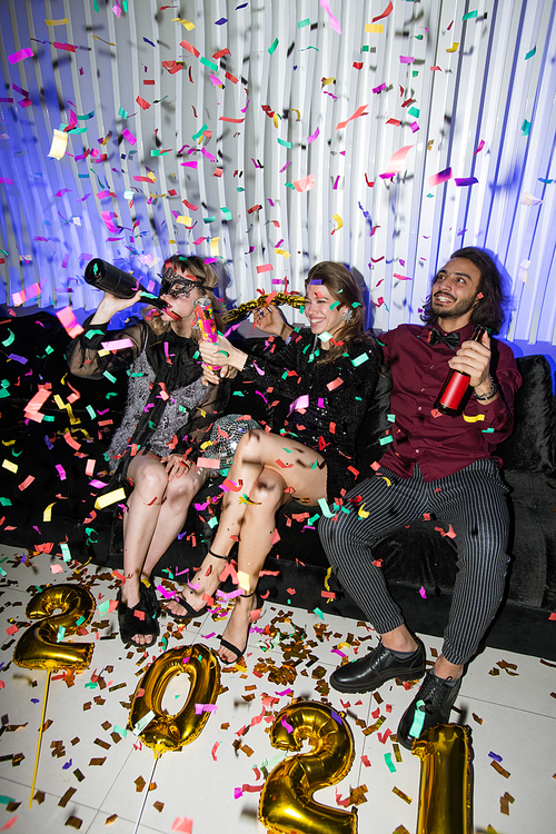 Group of happy young friends sitting on couch in the night club, relaxing and having beer under confetti