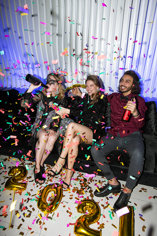 Young joyful friends with beer sitting on soft couch while having fun during new year party in the night club