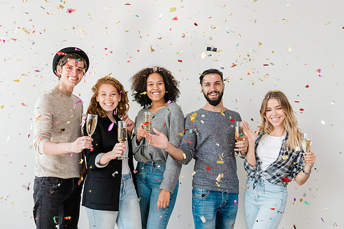 Young ecstatic multiracial friends cheering up with champagne in colorful confetti at home party