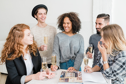 Laughing friends with flutes of champagne looking at one of girls telling funny stuff by table at home party