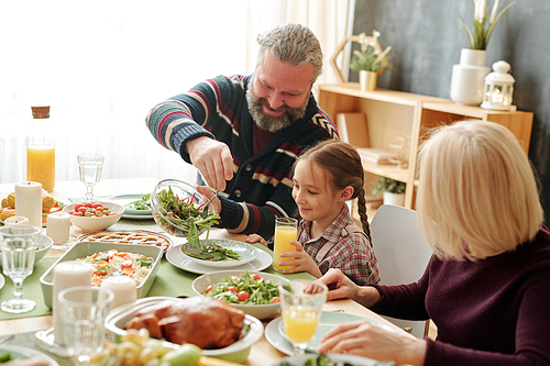 Happy grandpa giving salad to his granddaughter by festive table during Thanksgiving or Christmas family dinner at home