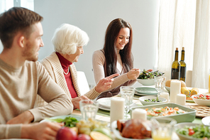 Happy young brunette woman putting salad on plate of grandma sitting next to her by festive table served for family dinner