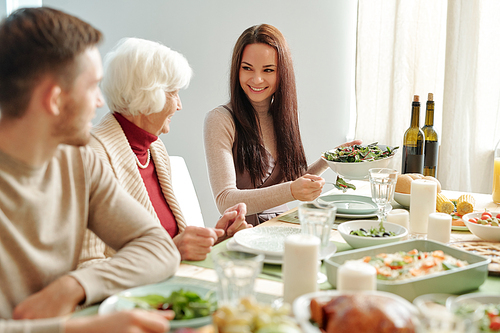 Smiling young woman putting salad on plate of grandma while looking at her by festive table during family dinner