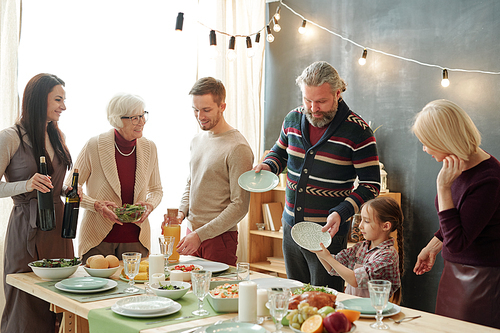 Family of five standing by served festive table on holiday, taking plates for snacks and preparing drinks before celebration