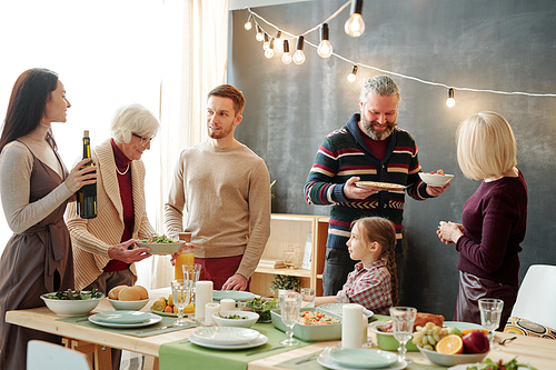 Mature and young couples chatting by festive table while preparing drinks and food for home festivity