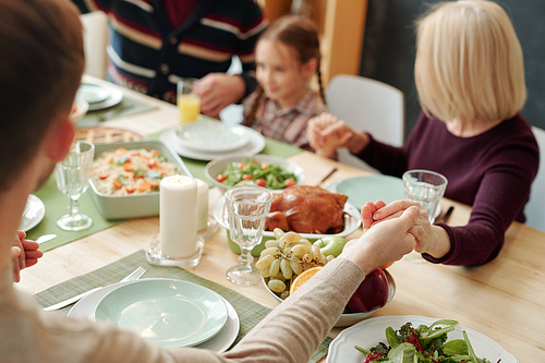 Hands of young man, mature blonde woman and the rest of family held by each other over served festive table during Thanksgiving pray