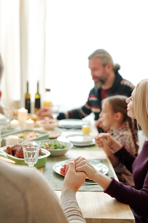 Young man, blonde female, her husband and little granddaughter holding by hands over festive table during Thanksgiving pray